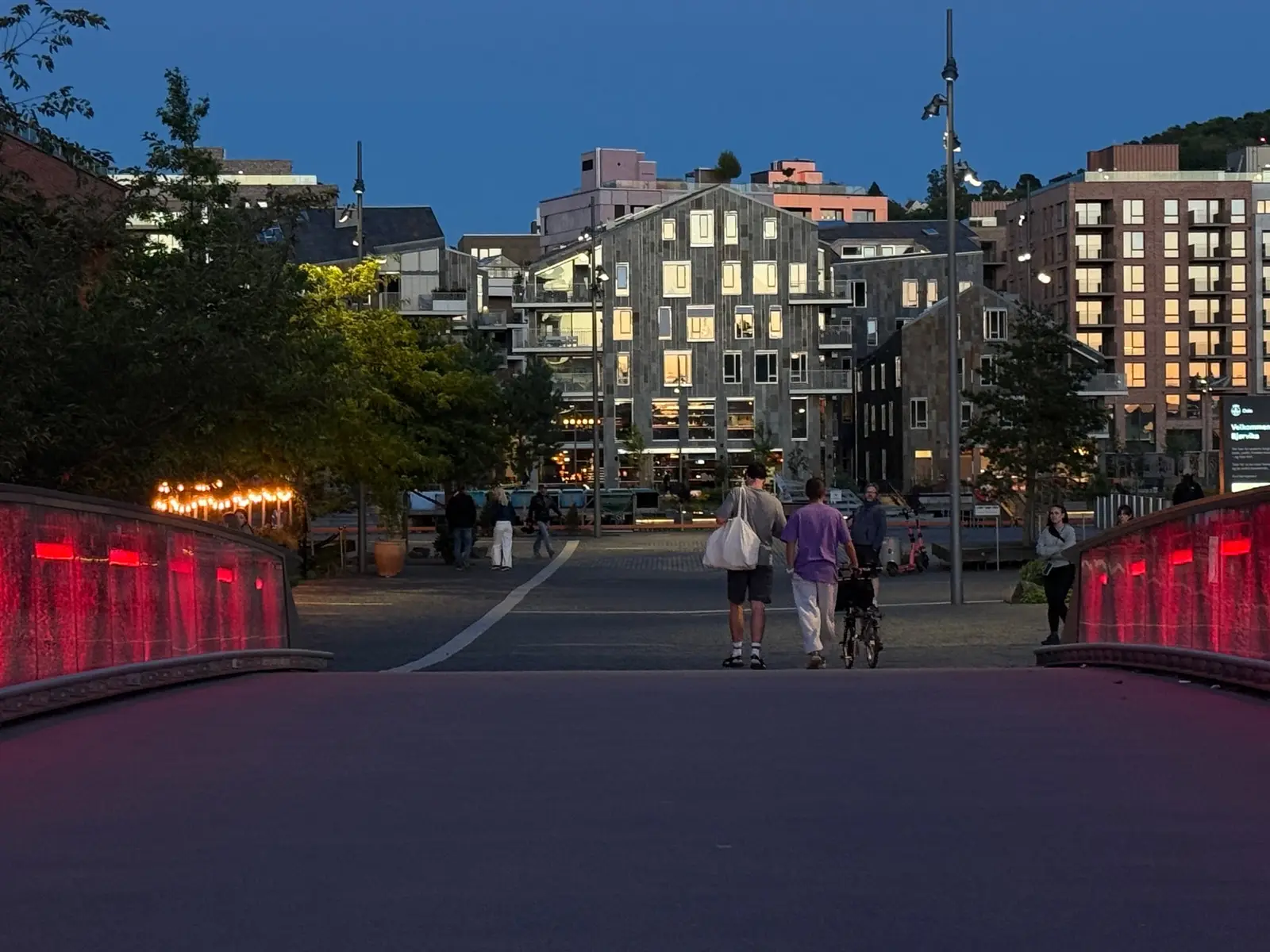Bispevika en sommerkveld: den røde brua, opplyste fasader og folk på vei over plassen.