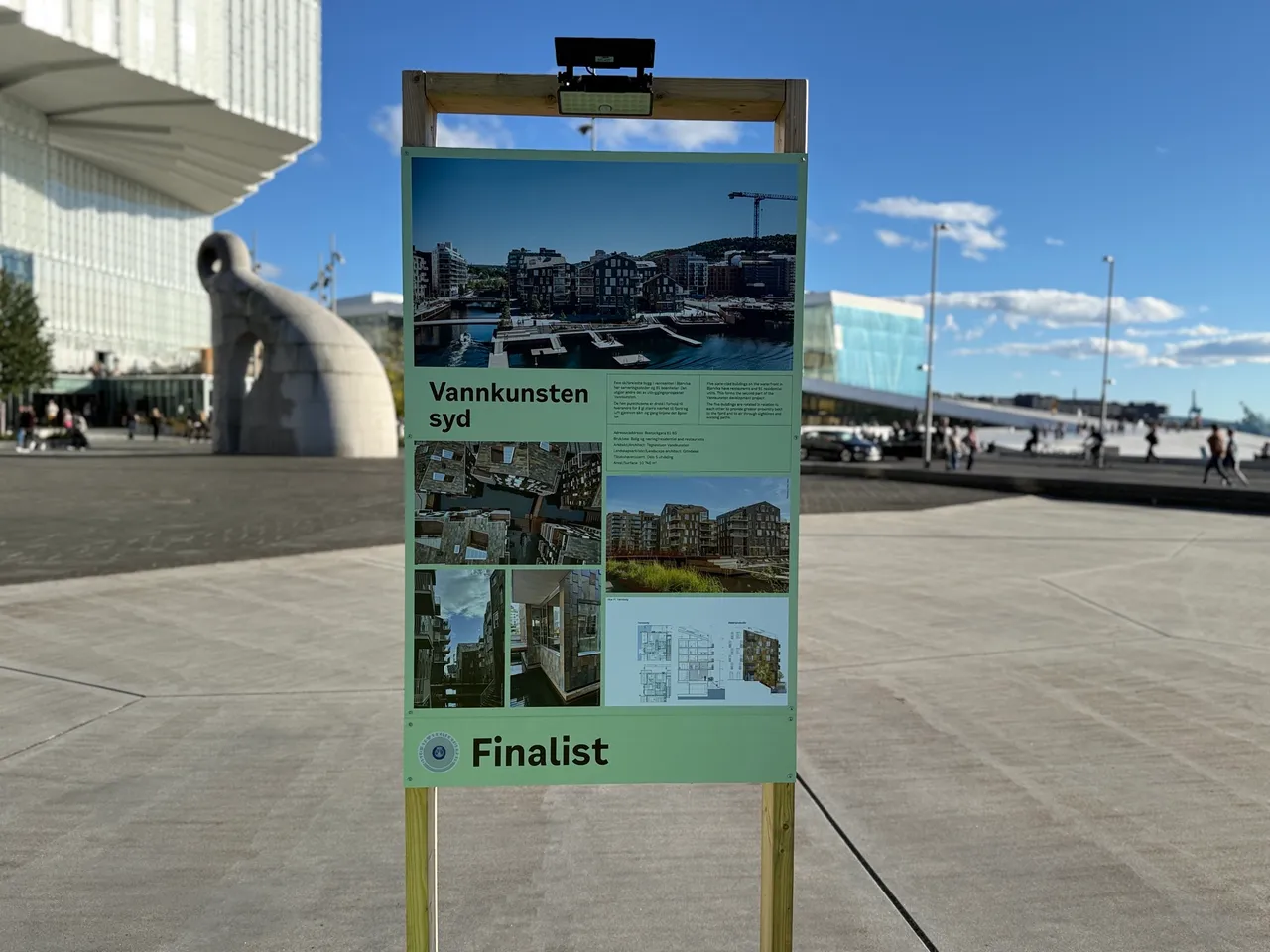 Utstillingsplakaten for Vannkunsten Syd står på plassen foran Deichman, med Operaen og fjorden bak. Nederst på plakaten står ordet Finalist.